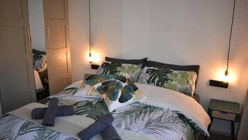 A neatly made bed with tropical leaf-patterned bedding, two bedside tables with wall-mounted lamps, and a mirror on the left wall.