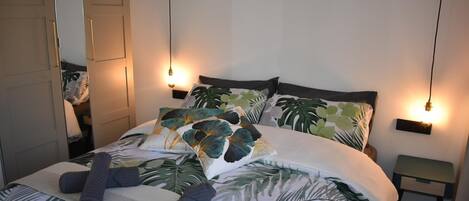 Una cama bien hecha con ropa de cama estampada con motivos de hojas tropicales, dos mesitas de noche con lámparas de pared y un espejo en la pared izquierda.