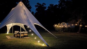 Una gran carpa blanca con un área de comedor en su interior, rodeada por un campo de césped y árboles.