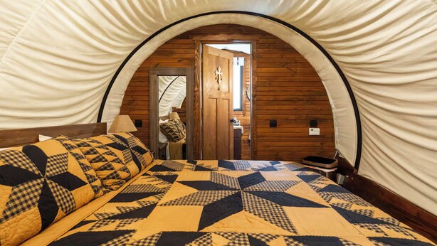 Una habitación acogedora con colcha de cuadros, paredes de madera y una puerta que da a otra estancia.