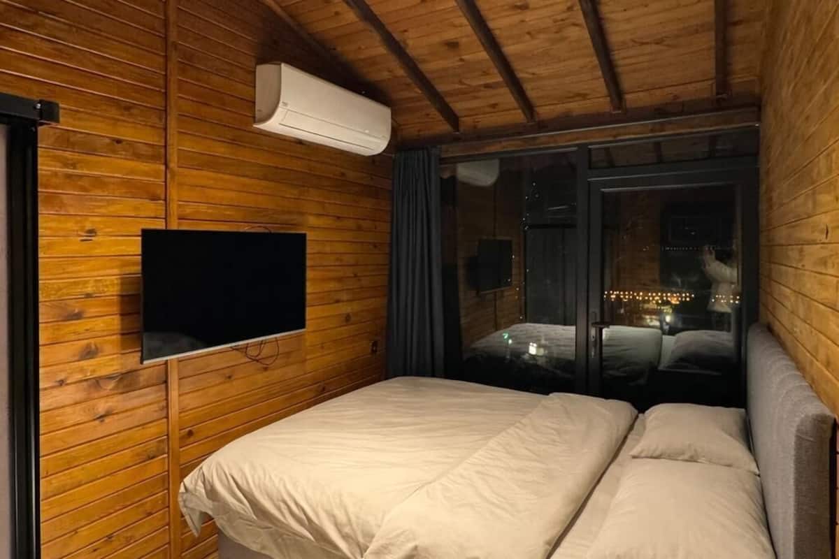 Una habitación acogedora con techo de madera, una cama con dos almohadas, un televisor en la pared y una ventana con cortinas.