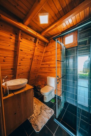 Baño de cabaña de madera con tocador de madera, lavamanos blanco, inodoro y mampara de vidrio para la ducha.
