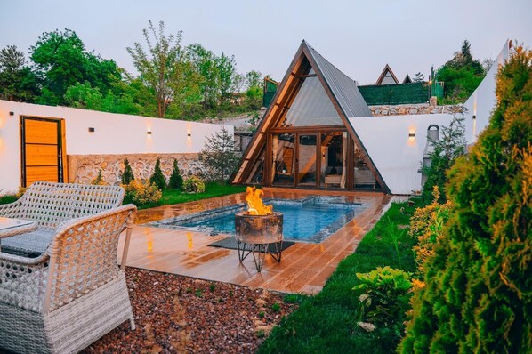 Un área de piscina moderna al aire libre con una estructura de vidrio triangular, mobiliario de mimbre y un brasero.