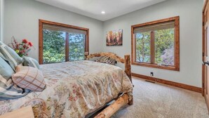 Un dormitorio con una cama grande, dos ventanas, un cuadro y vistas a árboles.