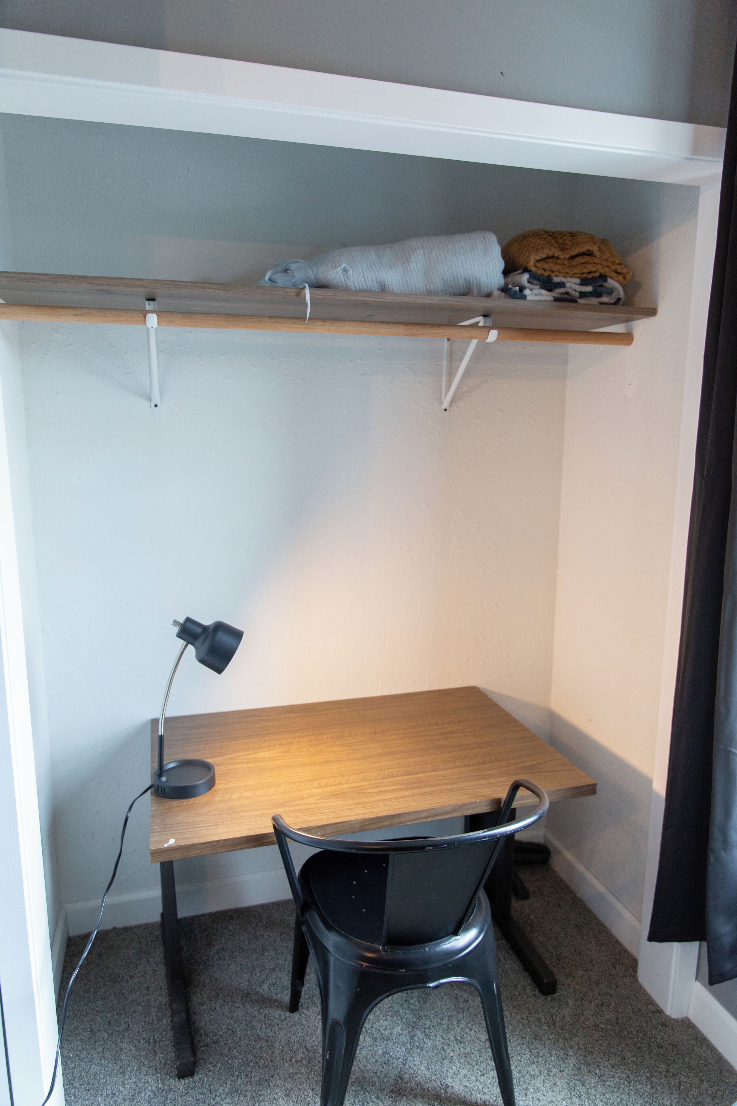 Une pièce de taille modeste, avec un bureau en bois, une chaise noire, une lampe de bureau noire et une étagère au-dessus du bureau.