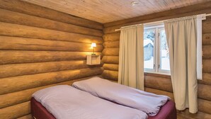 Une chambre rustique en rondins de bois, avec un lit, une table de chevet dotée d’une lampe et une fenêtre avec des rideaux.