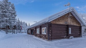 Une cabane en rondins de bois, avec un toit enneigé et une cheminée, entourée d’arbres couverts de neige et sous un ciel dégagé.