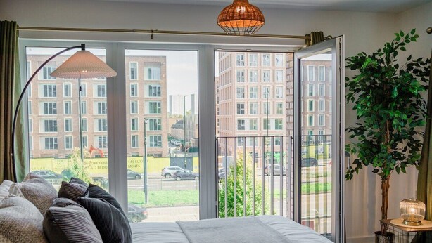 A bedroom with a large bed, a floor lamp, a potted plant, and a view of buildings through a glass door. - City Central | Free Parking | O2 | University (Birmingham)