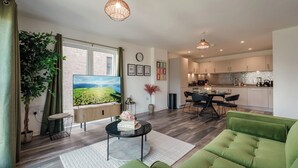 A modern living room with a green sofa, a coffee table, and a large TV. There is a dining area with a table and chairs, and a kitchen in the background. - City Central | Free Parking | O2 | University (Birmingham)