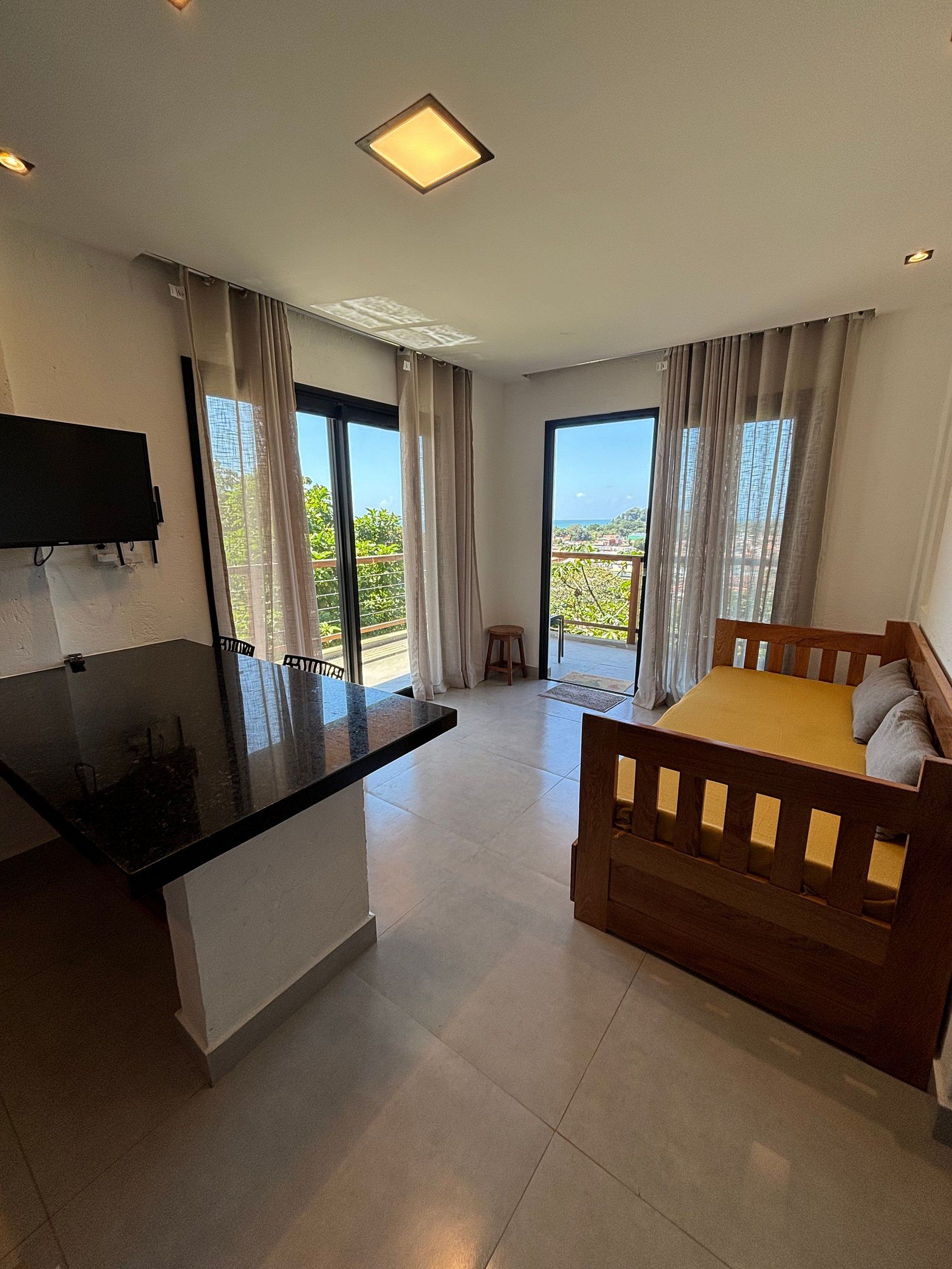A hotel room with a bed, a small stool, a flat-screen TV mounted on the wall, and a balcony with curtains.