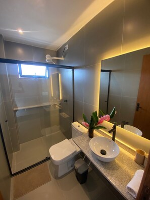A modern bathroom with a glass-enclosed shower, a white toilet, a sink with a vase of flowers, and a large mirror. - Villa Visgo Itacaré (Itacaré)