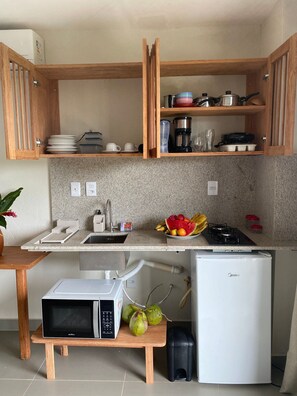 A compact kitchen with wooden cabinets, a built-in microwave, a small refrigerator, and a countertop with a fruit bowl. - Villa Visgo Itacaré (Itacaré)