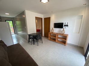 A living room with a sofa, a dining table with chairs, a TV on a stand, and a door leading to another room. - Villa Visgo Itacaré (Itacaré)