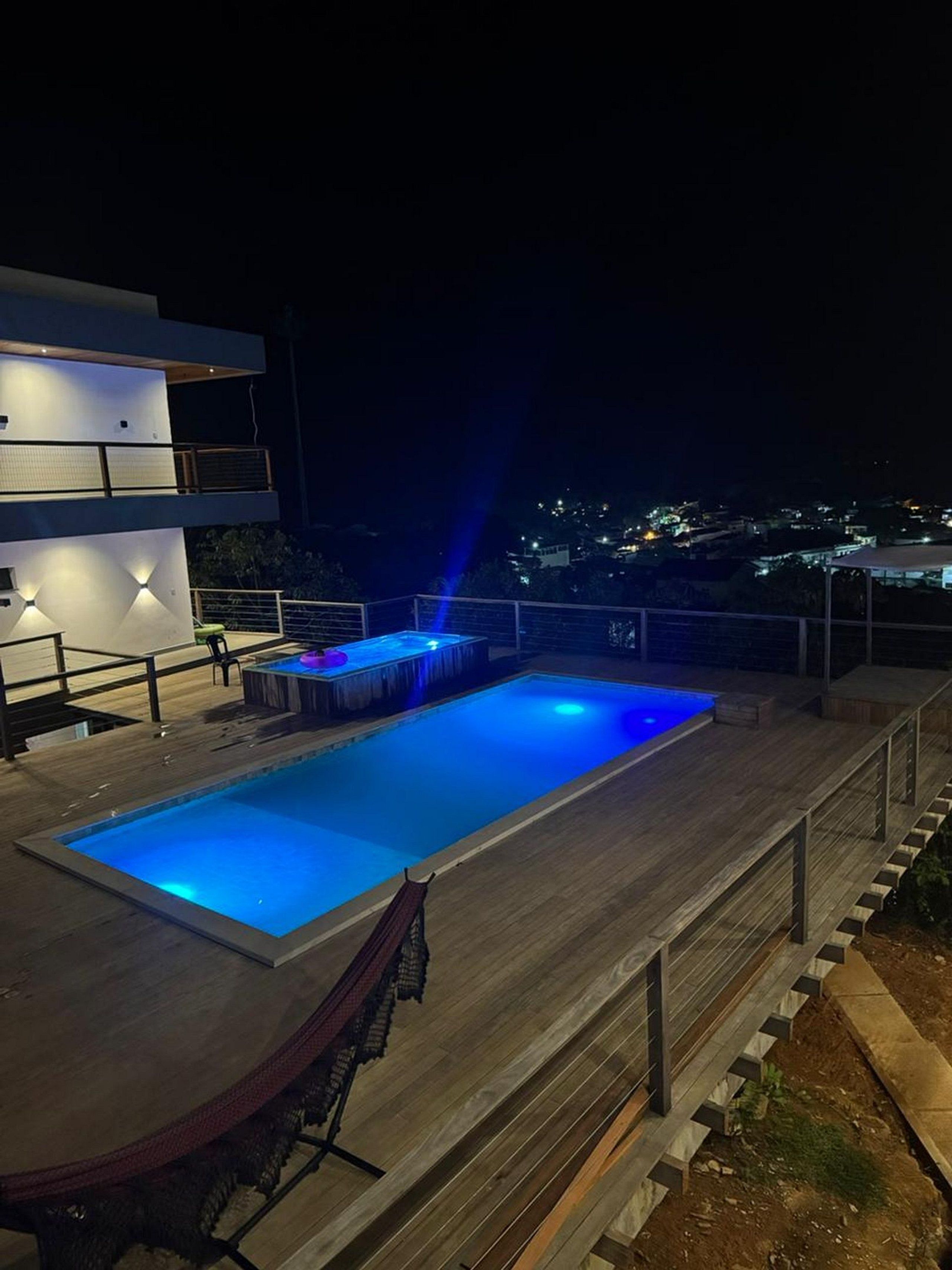 A swimming pool with a wooden deck, a modern building with illuminated walls, and a view of a distant town at night.
