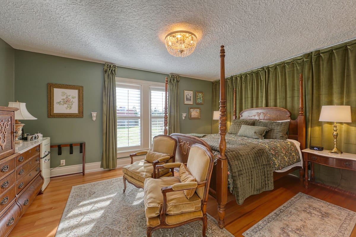 A bedroom with a four-poster bed, a wooden dresser, a chair, and a nightstand with a lamp.