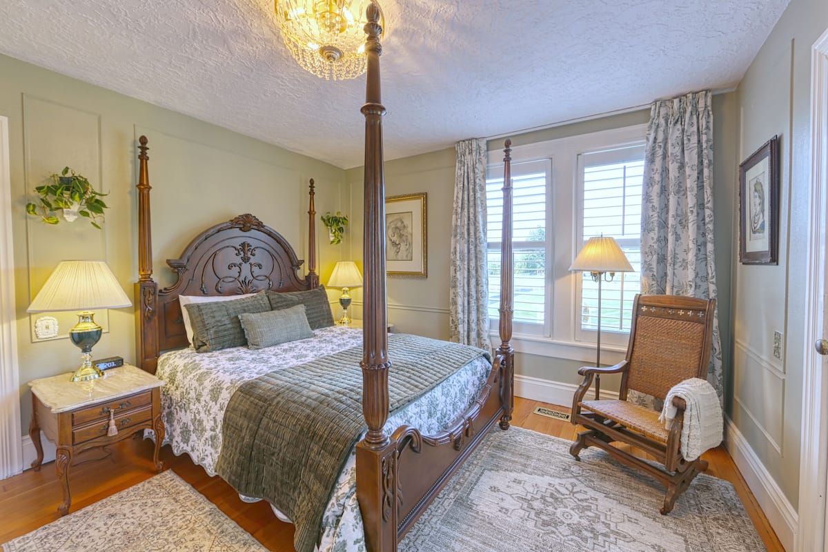 A bedroom with a four-poster bed, two bedside tables with lamps, a chair, and a large window with curtains.
