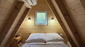 Ett mysigt sovrum på vinden med ett sluttande tak, ett fönster med utsikt över grönskan, en säng med ett täcke och två vägglampor.