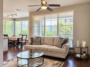 A modern living room with a beige sofa, patterned cushions, a glass coffee table, and a dining area with a table and chairs. -  2BR Corporate Stay Near UT Southwestern & Love Field (Dallas)