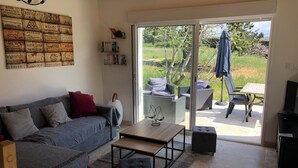 A living room with a grey sofa, a coffee table, and a bookshelf. There is a sliding glass door leading to an outdoor patio area with wicker furniture and a TV. - Charming cottage (Monteux)