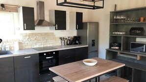 A modern kitchen with dark cabinets, a built-in oven, a stainless steel refrigerator, and a wooden dining table with a bowl. - Charming cottage (Monteux)