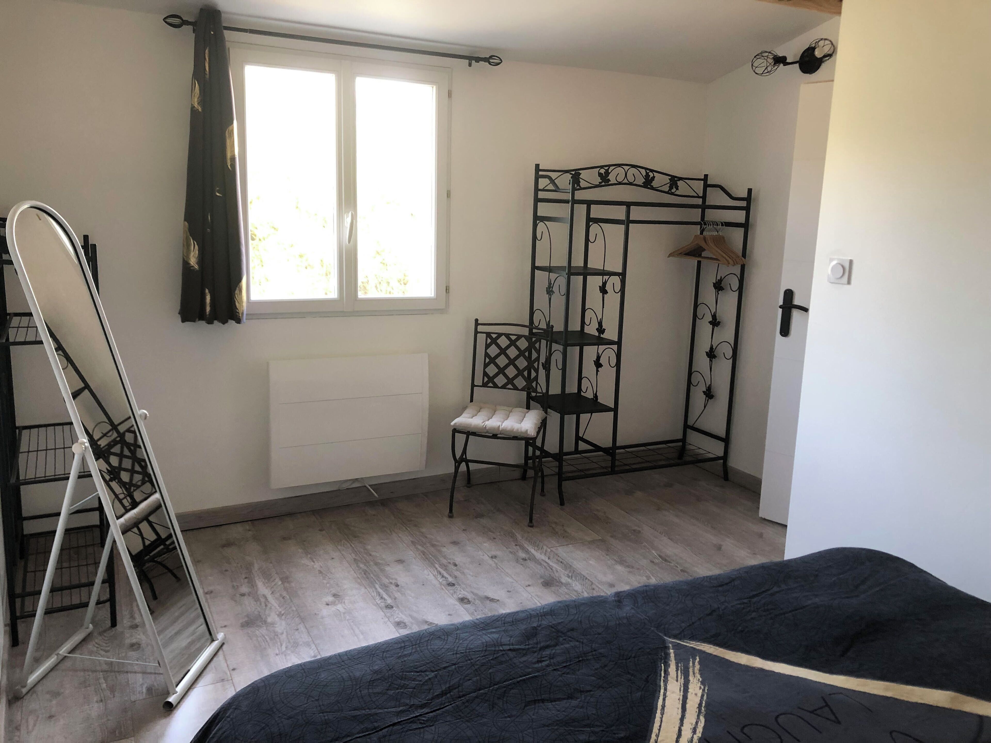 A room with a bed, a radiator, a window with curtains, a metal shelf unit, and a hanging rack.