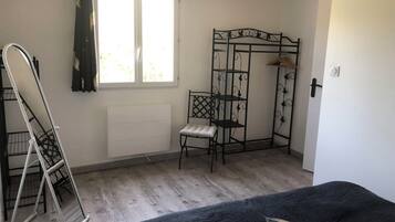 A room with a bed, a radiator, a window with curtains, a metal shelf unit, and a hanging rack.