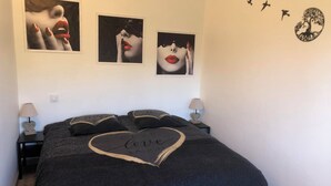 A bedroom with a black bedspread, two bedside tables with lamps, and three wall art pieces depicting abstract faces. - Charming cottage (Monteux)
