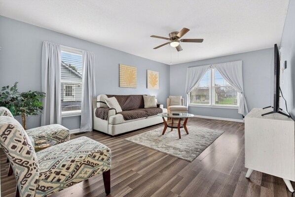 A living room with a sofa, armchair, and coffee table, a ceiling fan, and large windows with curtains. - Eagles Rest Home • Sleeps 7 • Minutes to Army Base (Oak Grove)