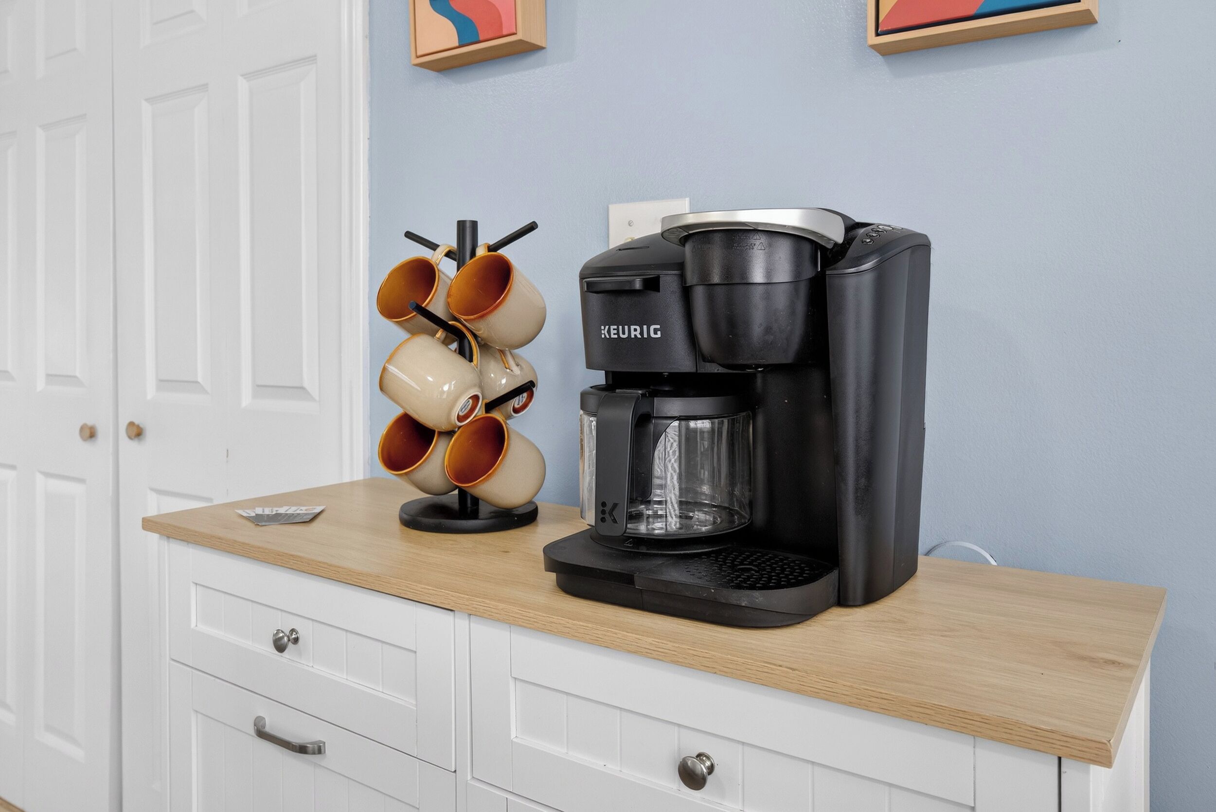Una cafetera sobre una encimera de madera, con un portal tazas para café y un mueble blanco debajo.