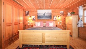 Une chambre à coucher aménagée dans une cabane en bois, avec un lit, des murs et un plafond en bois, et ornée d’un grand tableau représentant un cerf au-dessus du lit, ainsi que de plusieurs autres tableaux plus petits sur le thème du cerf.