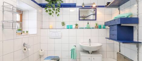 Ein Badezimmer mit weißen Fliesen, einer Toilette mit Sitzgarnitur im Stranddesign, einem Waschbecken, einem Spiegel und Regalen mit Pflegeprodukten.