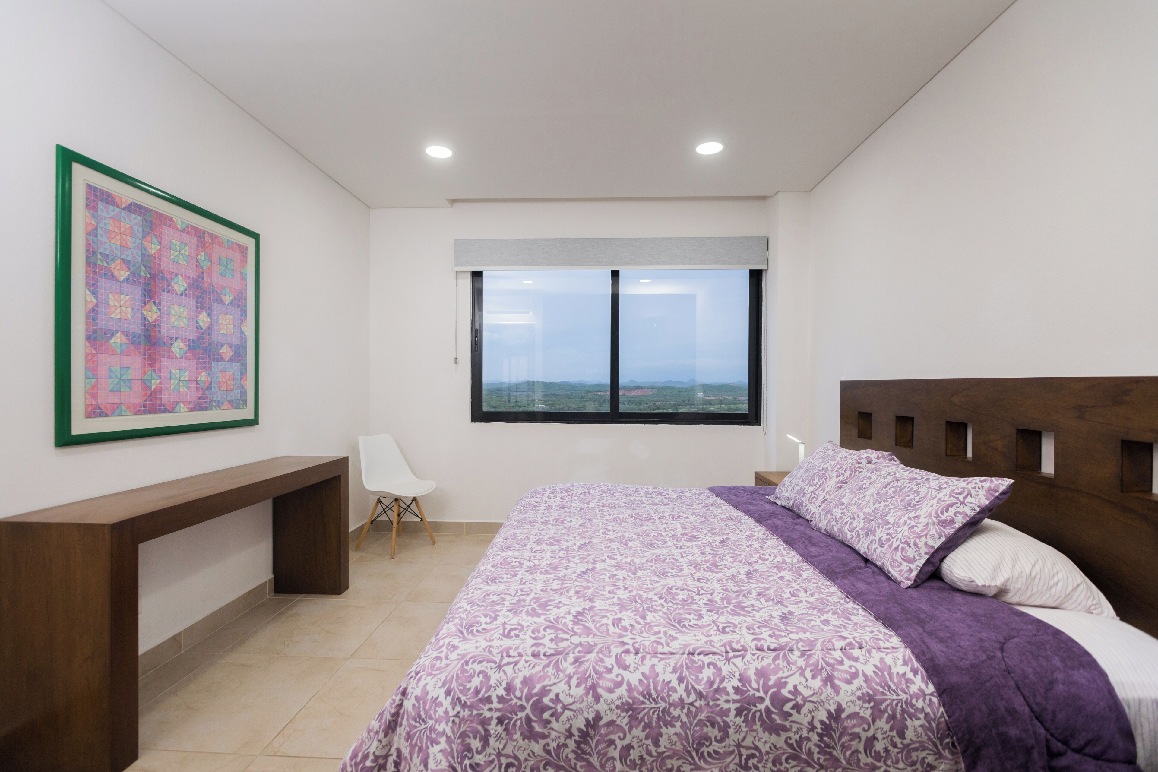 Een slaapkamer met een groot bed, een bureau, een stoel, een raam met uitzicht en een kunstwerk in een lijst aan de muur.