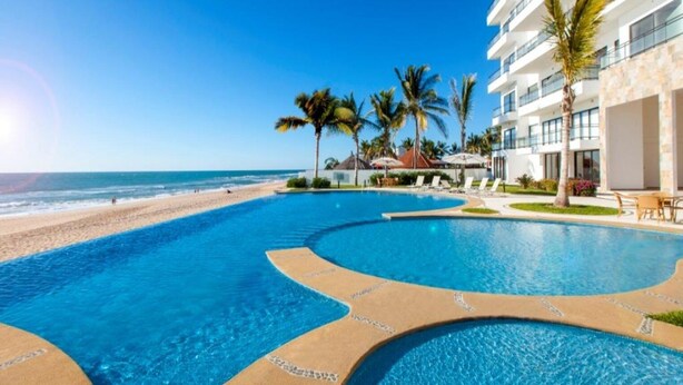 A swimming pool with a beach view, palm trees, and a building with balconies. - 3 bedroom high end beach front condo with amazing view (Mazatlán)