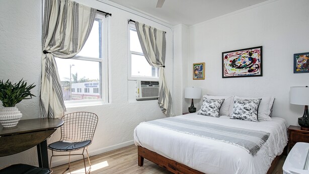 Une chambre à coucher avec un lit en bois, un bureau avec une chaise, une plante en pot et deux fenêtres avec des rideaux.