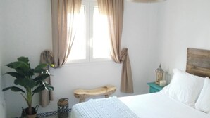 Ein Schlafzimmer mit einem Bett, einer hölzernen Bank, einem Blumentopf, einem Nachttisch mit einer Laterne und einem Fenster mit Vorhängen.