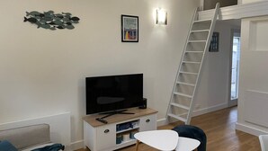 A modern living room with a flat-screen TV on a wooden TV stand, a white wall-mounted shelf with decorative items, a white ladder, and a staircase leading to an upper level. - Vent de Mer - House with a pool for 8 people (Bretignolles-sur-Mer)