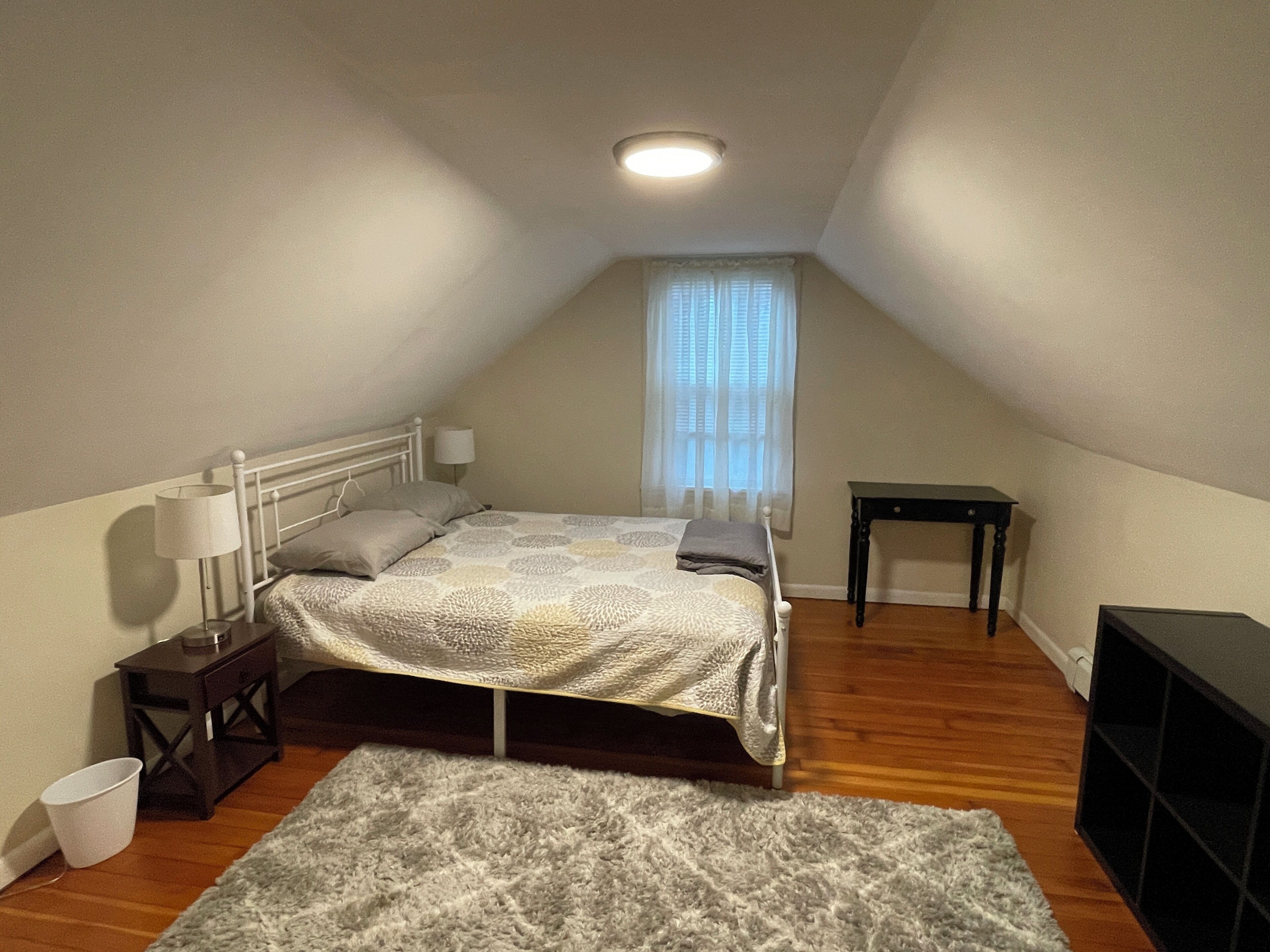Ein kleines, helles Schlafzimmer im Dachgeschoss mit einem Bett, Nachttischen, einem kleinen Tischchen, einem Fenster mit durchsichtigen Vorhängen und Holzboden.