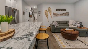 A modern kitchen with a marble countertop and wooden stools, a grey sofa, and a 'FAMILY' wall decoration. - Apex Slopeside Escape ~ Ski in/out with Hot tub (Keremeos)