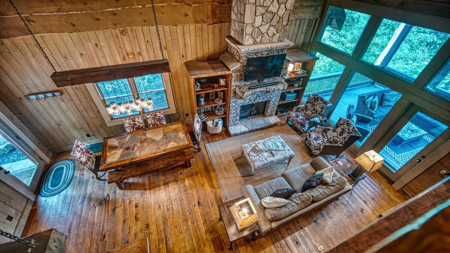 A cozy cabin interior with a stone fireplace, wooden furniture, and large windows offering views of the outdoors.