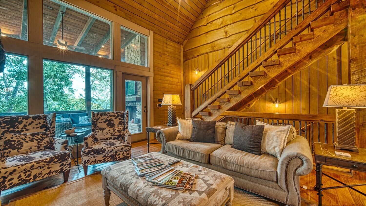 A cozy cabin interior with a wooden staircase, large windows, and a comfortable seating area.