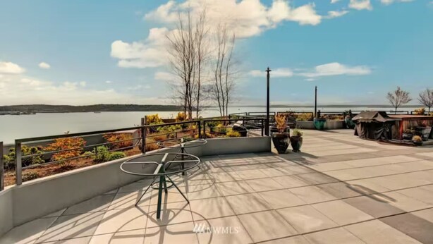 Terraço com vista para um corpo de água, cadeiras de metal, plantas em vasos e uma pequena área de estar ao ar livre.