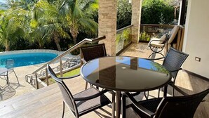 Et bord med glasplade og fire stole i et overdækket udendørsområde med swimmingpool og palmer i baggrunden.