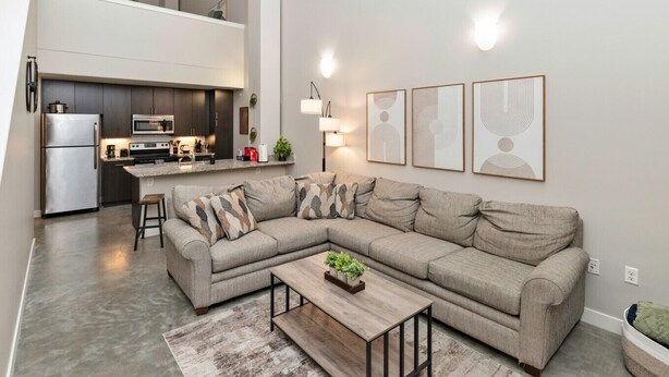 Een moderne woonkamer met een hoekbank, een houten salontafel, en een keuken met een koelkast, magnetron en fornuis.