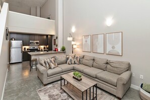 Una sala de estar moderna con un sofá modular, una mesa de centro de madera y una cocina equipada con refrigerador, microondas y estufa.