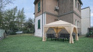 Een overdekt terras met tafels en stoelen, een gazon en een gebouw met een beige gevel en groene luiken op de achtergrond.