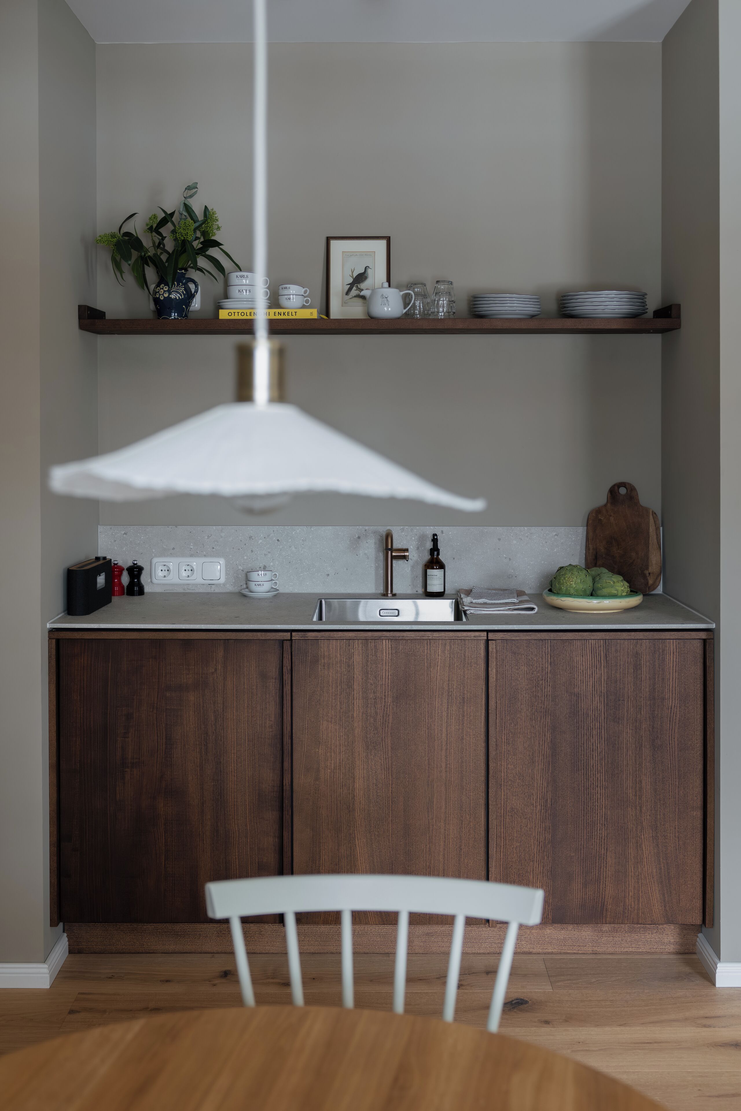 Une cuisine avec des meubles de rangement en bois, une chaise blanche, un luminaire suspendu et une étagère contenant des ustensiles de cuisine.