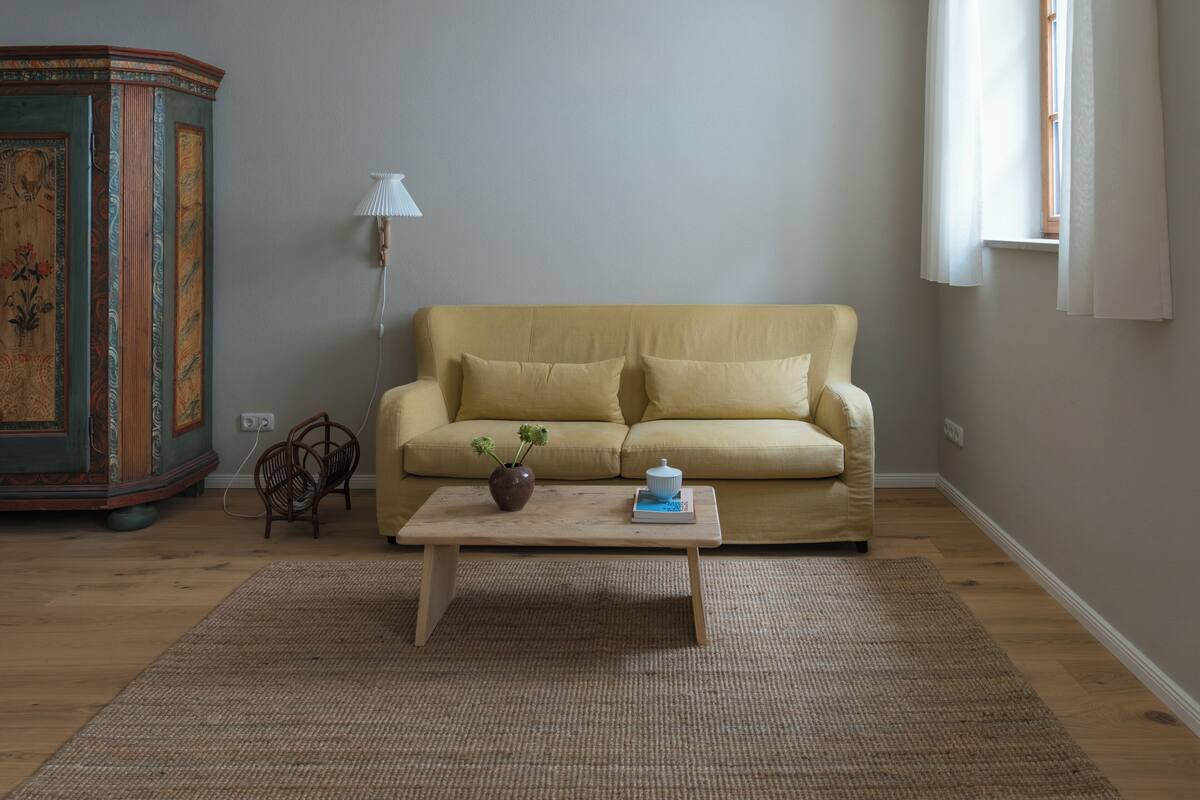 A living room with a beige sofa, a wooden coffee table, a decorative cabinet, a lamp, and a window with curtains.