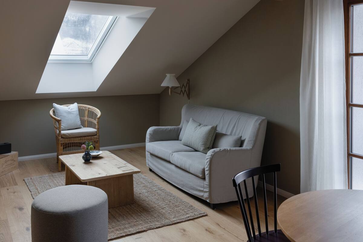 A cozy loft bedroom with a sloped ceiling, a grey sofa, a wooden coffee table, a wicker chair, and a small round table.