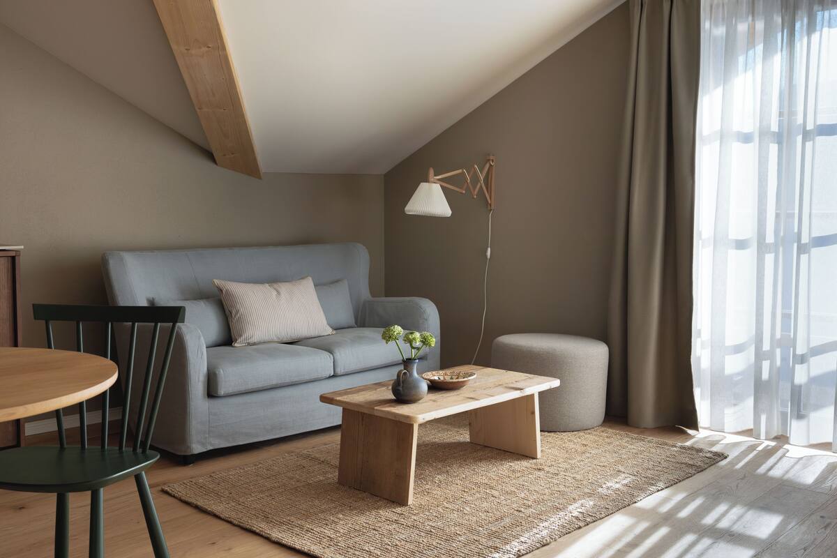 A modern living room with a grey sofa, a wooden coffee table, a floor lamp, and a large window with sheer curtains.