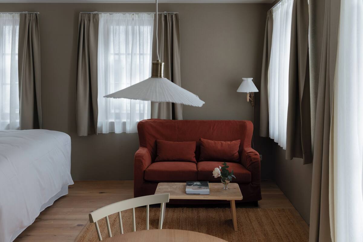 A room with a bed, a red sofa, a wooden coffee table with a book and a vase, a hanging lamp, and sheer curtains.